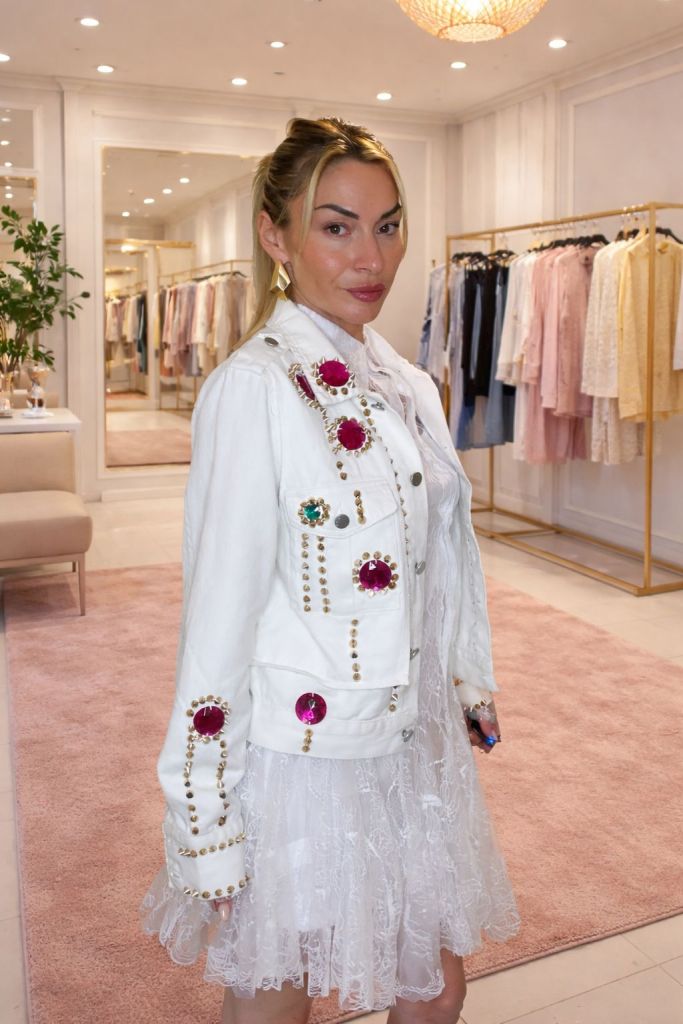Una donna in un negozio di abbigliamento indossa una giacca di jeans bianca decorata con gemme colorate e un vestito bianco in pizzo. Sullo sfondo si vedono abiti appesi e una decorazione elegante.