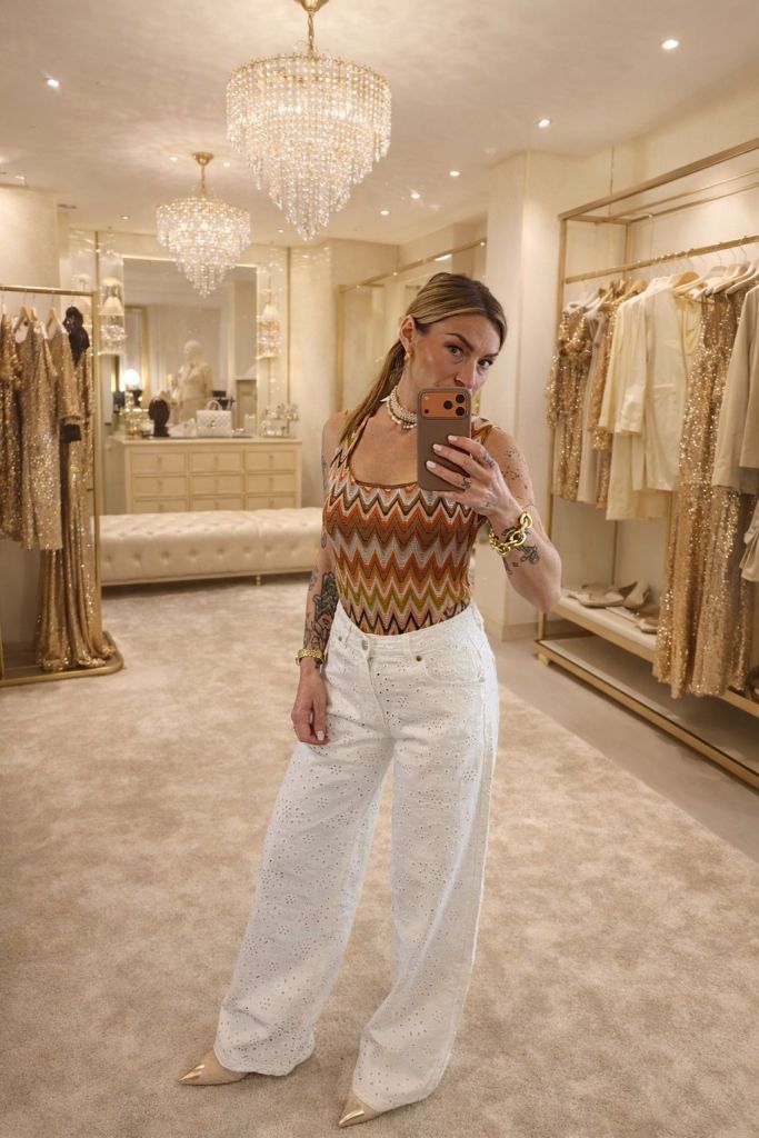 Una donna in un grande spogliatoio elegante, indossa un top a strisce e pantaloni bianchi. Sullo sfondo, ci sono abiti scintillanti e lampadari pendenti.