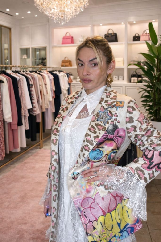 Donna in un boutique elegante, indossa un cappotto decorato con un motivo leopardato e grafiche colorate, mentre tiene la mano su un fianco.