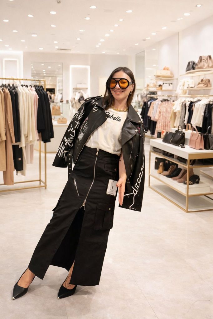 Una giovane donna sorridente in un negozio di moda, indossa una giacca di pelle nera con scritte artistiche e occhiali da sole arancioni. La sua outfit comprende una maglietta bianca e una gonna nera lunga con zip. Sullo sfondo, ci sono scaffali con abbigliamento e accessori.