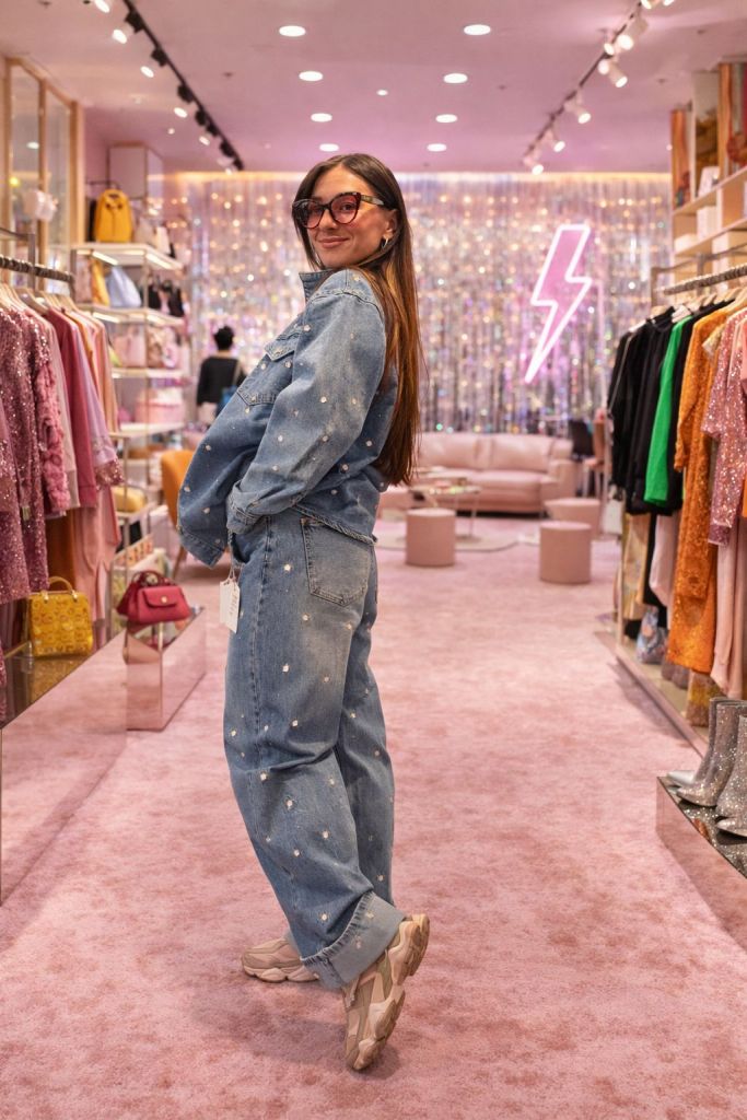 Una giovane donna in jeans con decorazioni e occhiali grandi, si gira sorridendo in un negozio di moda con tappeto rosa e abbigliamento colorato intorno.