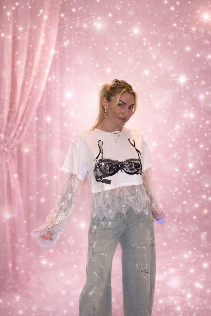 Una giovane donna con capelli biondi legati in una coda di cavallo indossa una maglietta bianca con un disegno di reggiseno nero e pantaloni larghi grigi, in un ambiente luminoso con sfondo rosa e scintille.