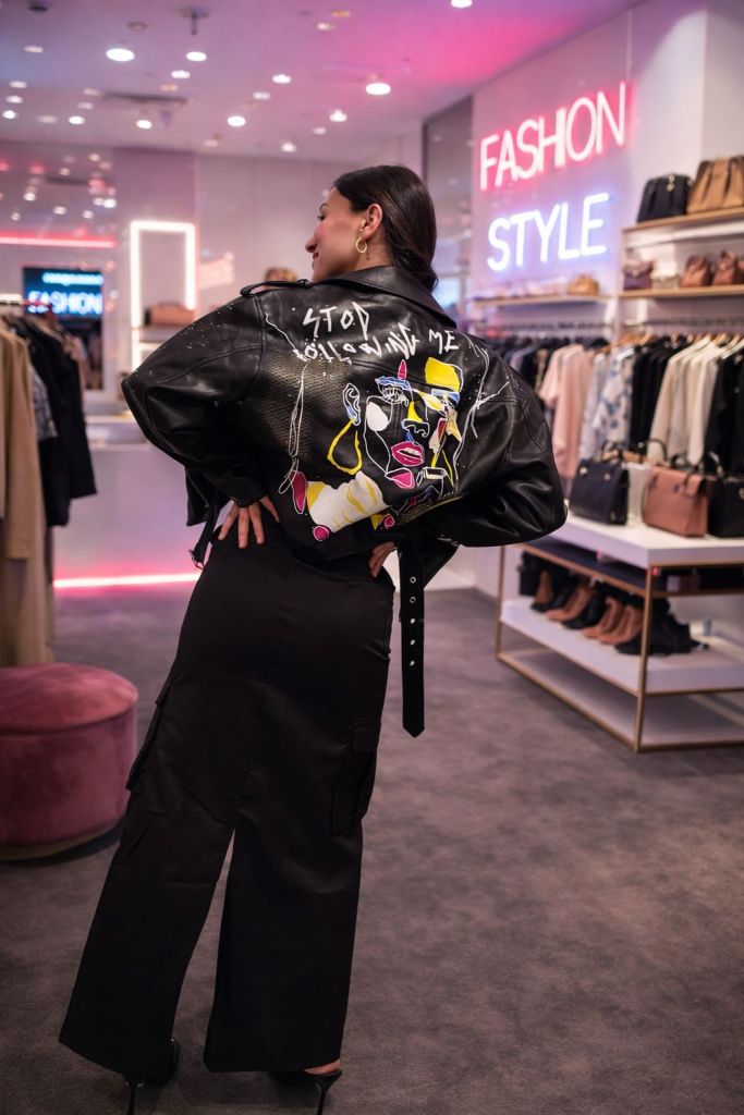 Una donna che indossa una giacca di pelle con un design artistico sul retro, si volta verso la telecamera in un negozio di moda. Sullo sfondo, una insegna luminosa con la scritta 'FASHION STYLE'.