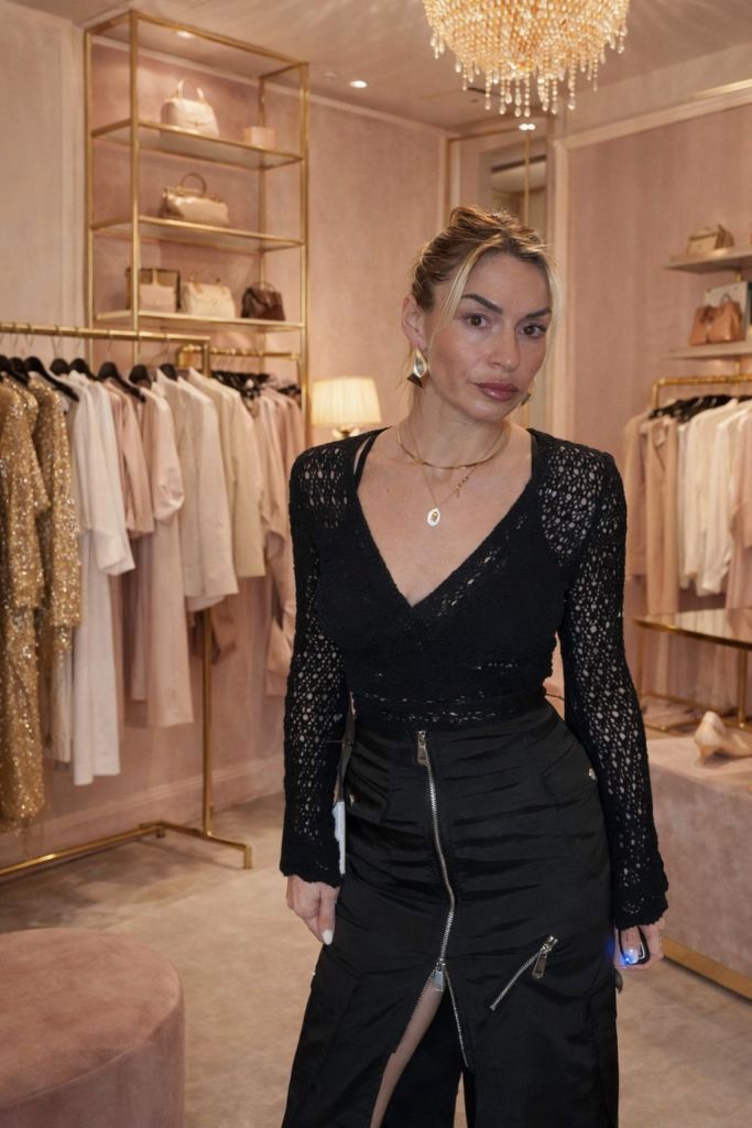 Una persona in abbigliamento elegante posa in un negozio di moda, circondata da vestiti e accessori in tonalità chiare, con un lampadario di cristallo sopra di lei.